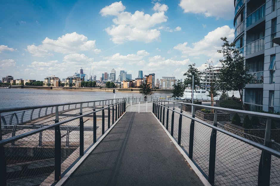 A wide view of a metal pedestrian walkway with protective fencing on both sides, leading from a residential or commercial building on the right toward a docked yacht and other boats moored along a riv