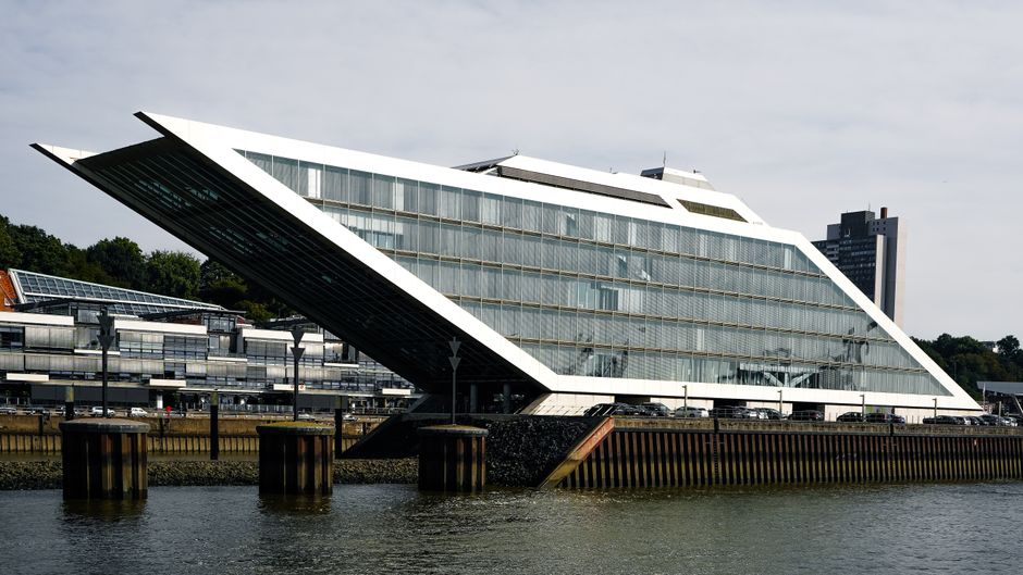 A daytime view of the Docklands waterfront showing modern high-rise office buildings and residential towers with glass facades reflecting sunlight. In the foreground, a concrete pier extends into the 