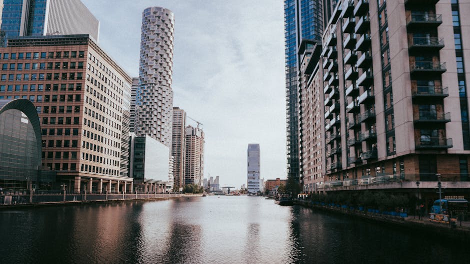 A cityscape featuring tall modern office buildings with reflective glass facades in the Docklands area, including the Canary Wharf district, under a cloudy sky. In the foreground, a red London rib-sty