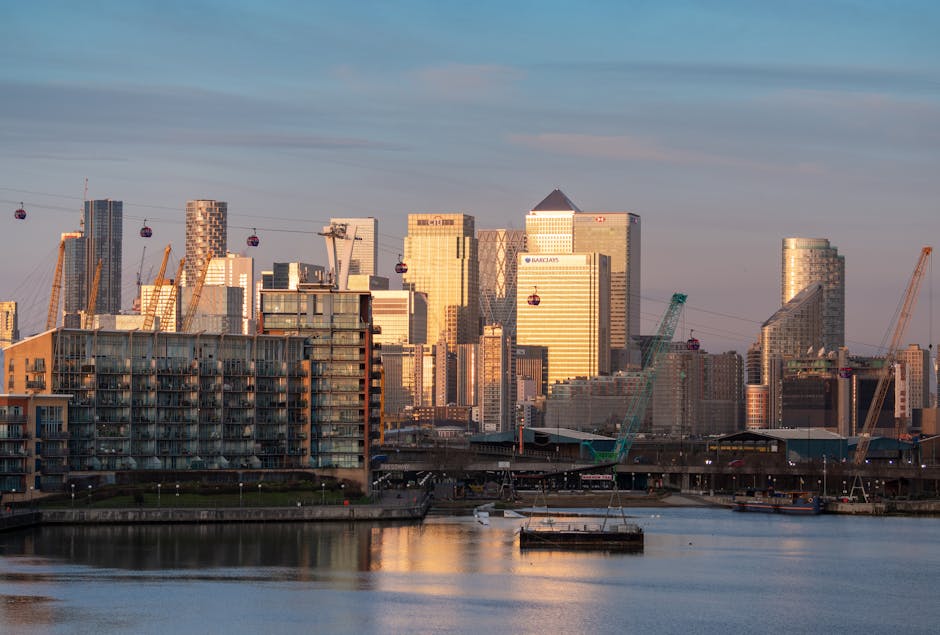 A daytime view of the Canary Wharf skyline in London, featuring several modern high-rise office buildings with glass facades, some displaying the Barclays logo. In the foreground, there is a river reflecting the buildings and the sky, with boats docked along the waterfront. Above, a cable car system with red cabins stretches across the skyline, indicating transport infrastructure associated with the area. Construction cranes are visible among the buildings, suggesting ongoing development. Part of a low-rise residential or commercial building with balconies is seen at the water's edge, along with a paved walkway or quay. The scene captures the urban environment typical of a busy commercial district, with natural lighting from the setting or rising sun casting a warm glow on the structures. This scene aligns with the context of home relocation and furniture transport, as depicted by the cityscape background associated with Docklands Movers' removals services.