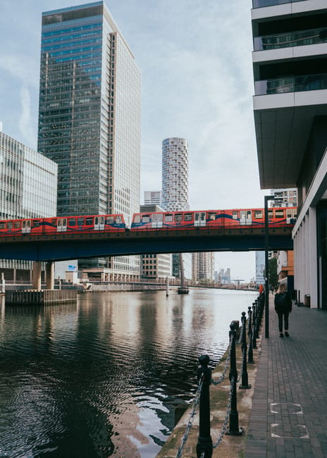 The image shows a modern urban scene in Canary Wharf with a red and white train crossing a bridge over a river, flanked by high-rise office buildings with glass facades. To the right, a person dressed in dark clothing is walking along a paved walkway beside the water, bordered by black metal bollards and chains. The river reflects the surrounding architecture, with some rippling on the surface. The sky is partly cloudy with soft-light conditions, highlighting the clean lines of the surrounding buildings and the bridge structure. This scene captures a typical day in the Docklands area during a home relocation or moving process, where nearby buildings and infrastructure such as train routes and pedestrian pathways facilitate efficient logistics and transportation, relevant to house removals and furniture transport services provided by Docklands Movers.