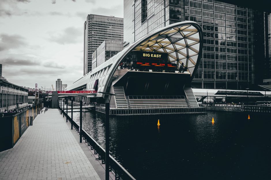 A daytime view of the Canary Wharf skyline in London, featuring several modern high-rise office buildings with glass facades, some displaying the Barclays logo. In the foreground, there is a river reflecting the buildings and the sky, with boats docked along the waterfront. Above, a cable car system with red cabins stretches across the skyline, indicating transport infrastructure associated with the area. Construction cranes are visible among the buildings, suggesting ongoing development. Part of a low-rise residential or commercial building with balconies is seen at the water's edge, along with a paved walkway or quay. The scene captures the urban environment typical of a busy commercial district, with natural lighting from the setting or rising sun casting a warm glow on the structures. This scene aligns with the context of home relocation and furniture transport, as depicted by the cityscape background associated with Docklands Movers' removals services.