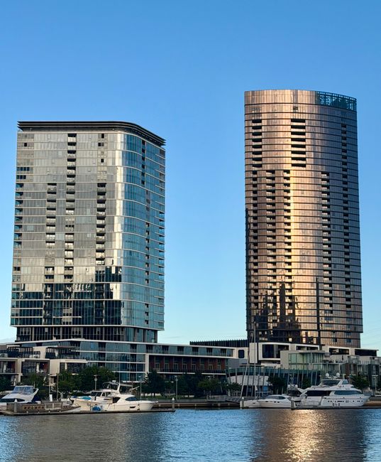 A daytime scene showing two modern high-rise buildings with glass facades situated along a waterfront. The building on the left has a rounded corner design with reflective windows, while the building on the right features a slightly curved structure with a mix of dark and light panels. Below the buildings, a marina is visible with several docked yachts and boats, with some boats partially obscured by the edge of the water. The sky is clear and blue, providing a bright backdrop for the urban landscape. This setting illustrates a waterfront property area often associated with city centre living or commercial developments, which is relevant to house removals and relocation services. Docklands Movers may assist with the safe transportation of belongings in such urban environments, including packing and load-in processes. The photograph emphasizes the mix of architecture and maritime environment, highlighting the logistical considerations involved in home relocation or furniture transport across property types in docklands areas.
