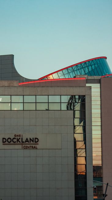 A modern multi-storey building with a predominantly glass facade and concrete sections, featuring a curved red roof element at the top. The building is located in the Docklands area, indicated by the sign reading 'One Dockland Central' visible on the lower part of the structure. The photograph is taken during daylight with clear skies in the background, highlighting the architectural design. While the image does not show moving or packing activities directly, it reflects the typical urban environment where house removals and furniture transport services by Docklands Movers might be required for local or transit-based relocations.