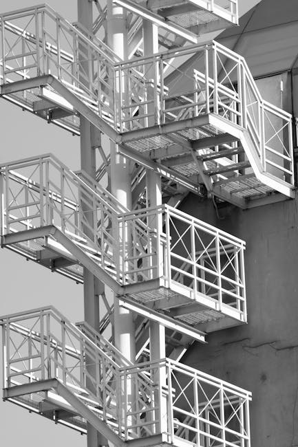 A black-and-white photograph showing an external emergency staircase attached to the side of a building, consisting of multiple landings connected by metal stairs with white railings. The staircases a